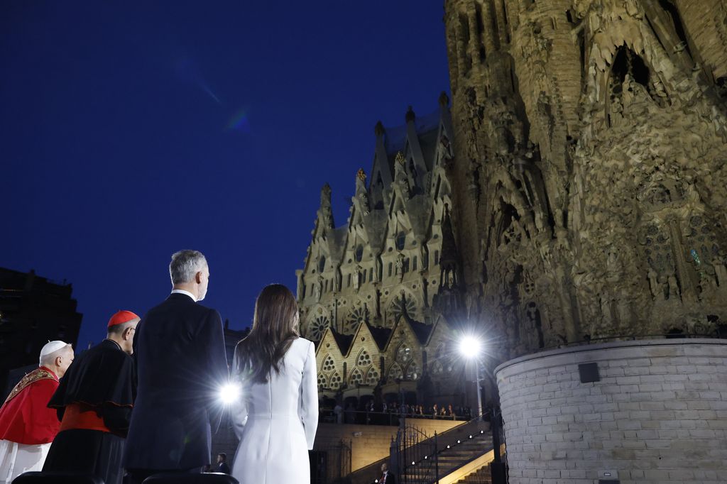 Tres personas, incluyendo dos clérigos, caminan frente a la Sagrada Familia al atardecer.