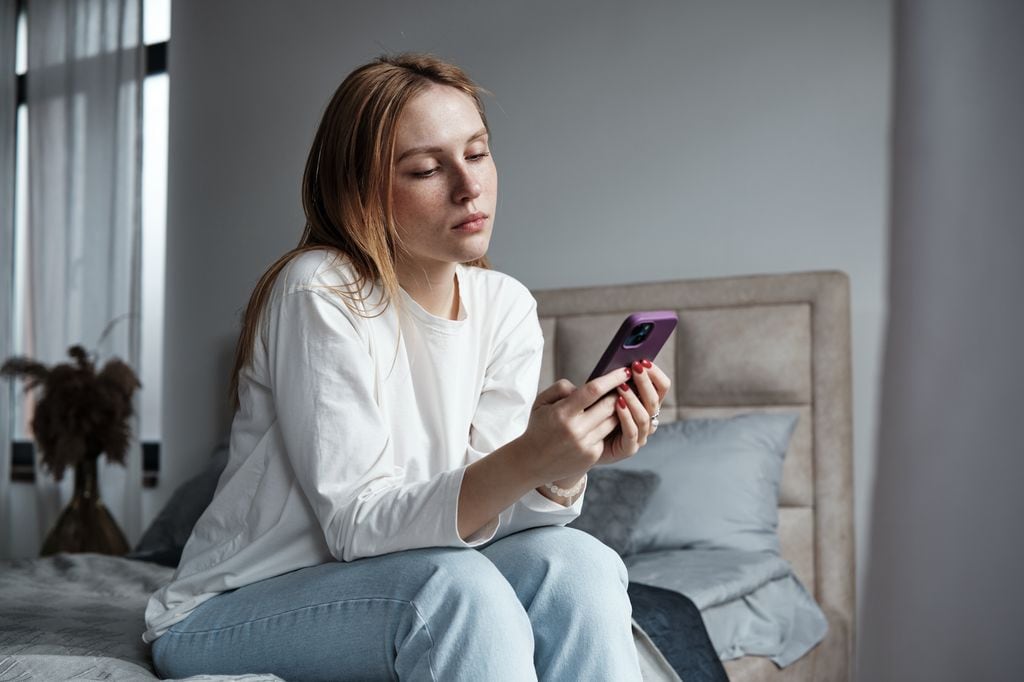 Chica triste sentada en la cama mirando el teléfono