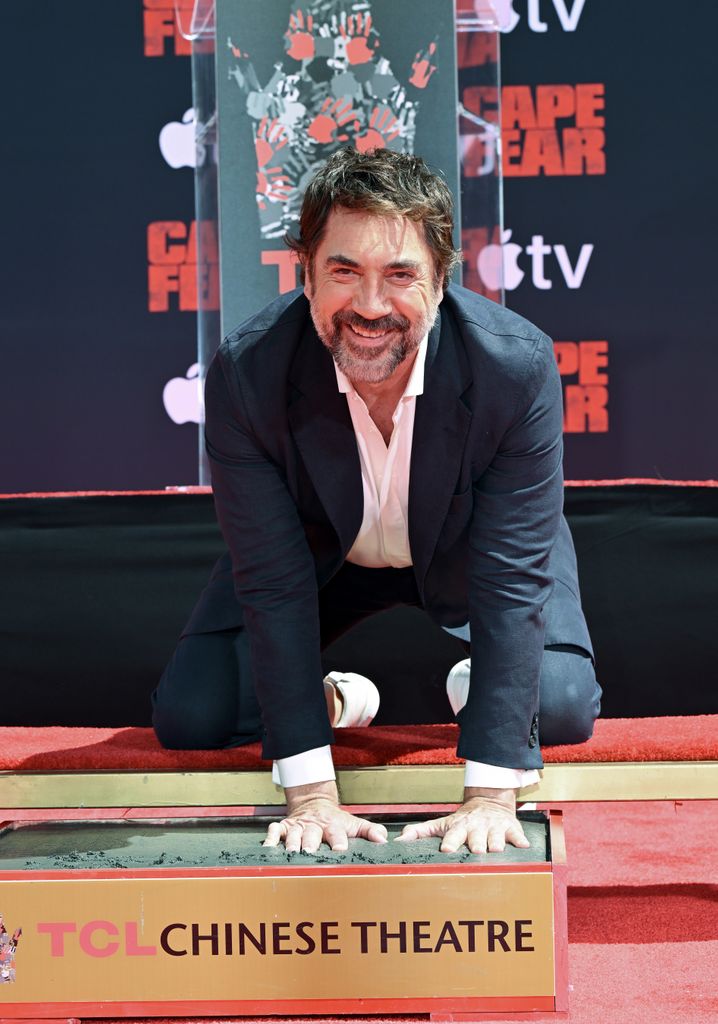 Javier Bardem en traje, arrodillado en una alfombra roja, sonriendo.