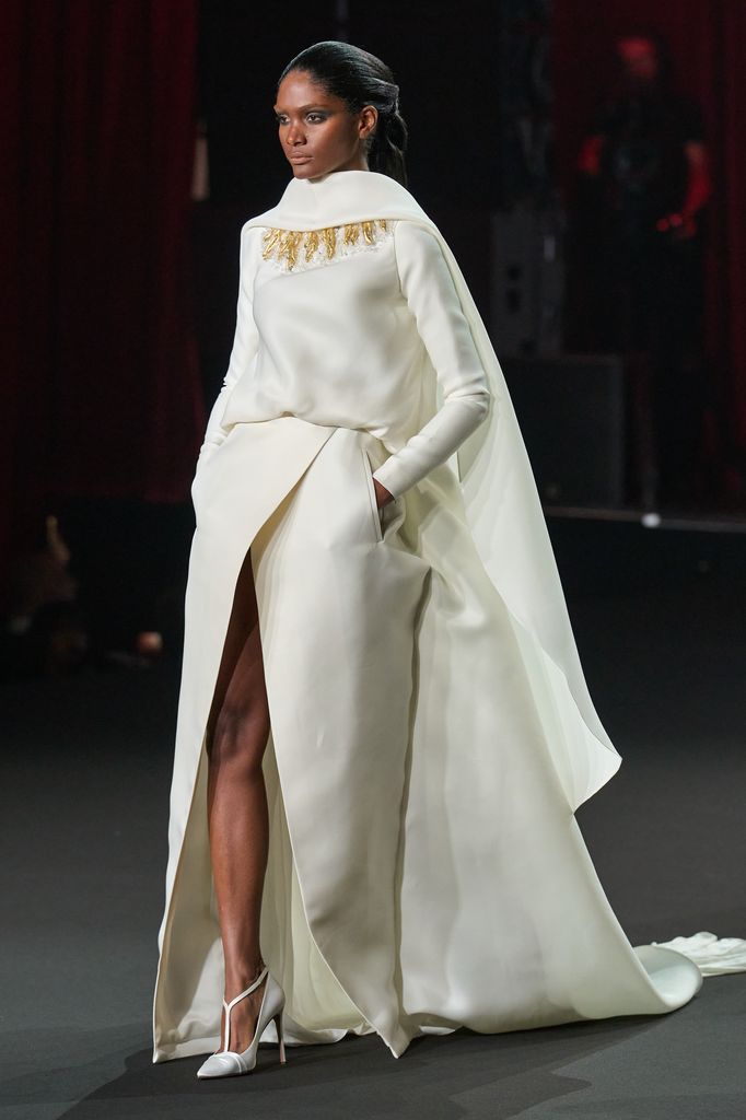 Modelo en pasarela con vestido blanco largo y capa dorada.