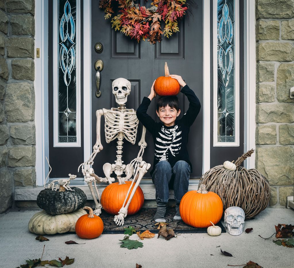 Un niño sonriente rodeado de calabazas y un esqueleto, celebrando Halloween en la puerta de su casa