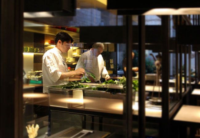 Dos chefs en uniforme blanco preparan alimentos en una cocina moderna.
