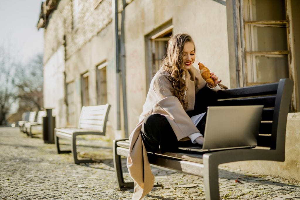 mujer comiéndose un croissant, mientras está sentada en un banco mirando su ordenador