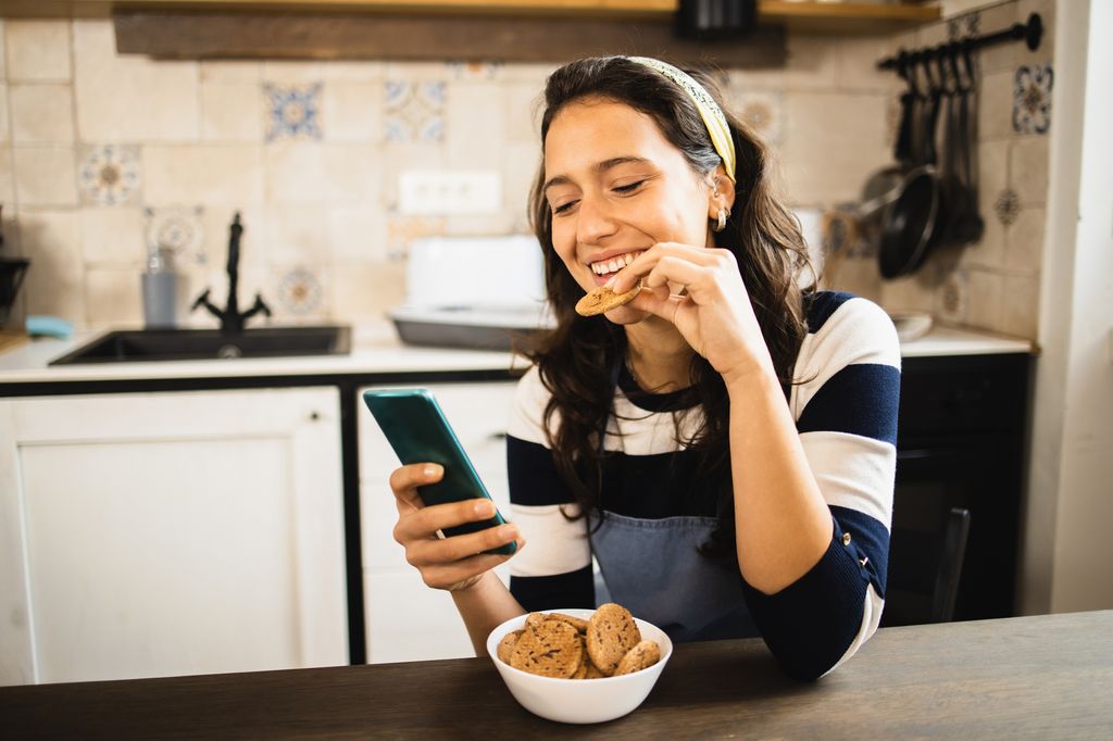 mujer comiendo una galleta en la cocina mientras mira su móvil