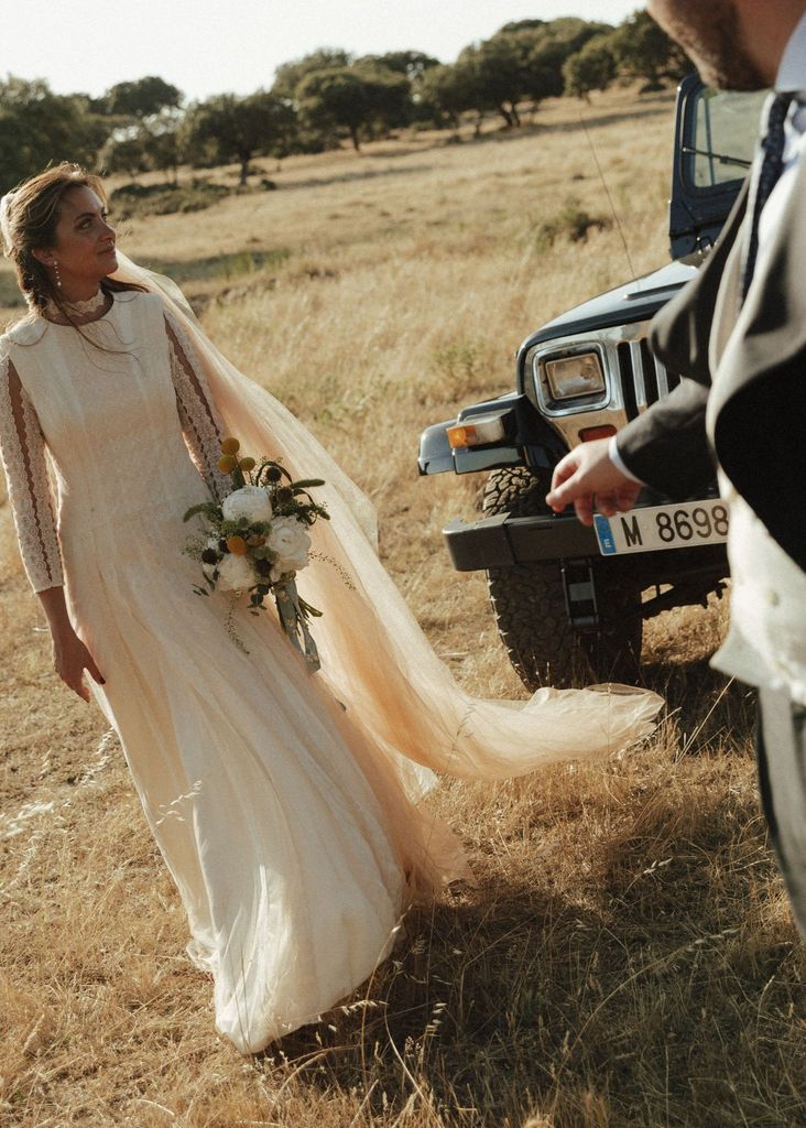 Novia en vestido blanco con velo y ramo, junto a un hombre en traje cerca de un jeep.