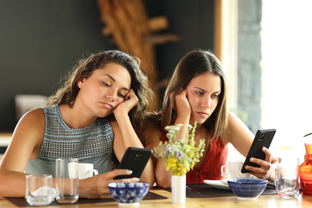 dos chicas jóvenes aburridas en un restaurante mirando sus móviles