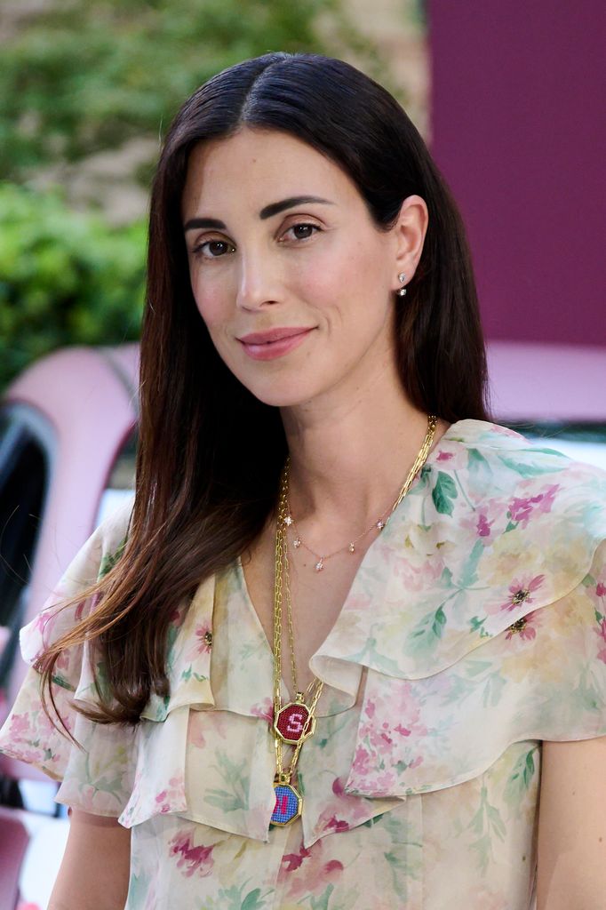 Mujer con cabello largo, blusa floral, y collar con dijes, sonriendo.
