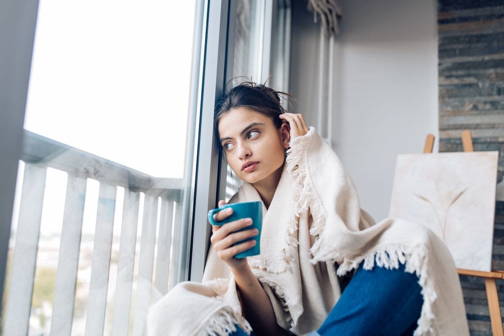 mujer tomando un té cubierta con una manta, mirando por la ventana