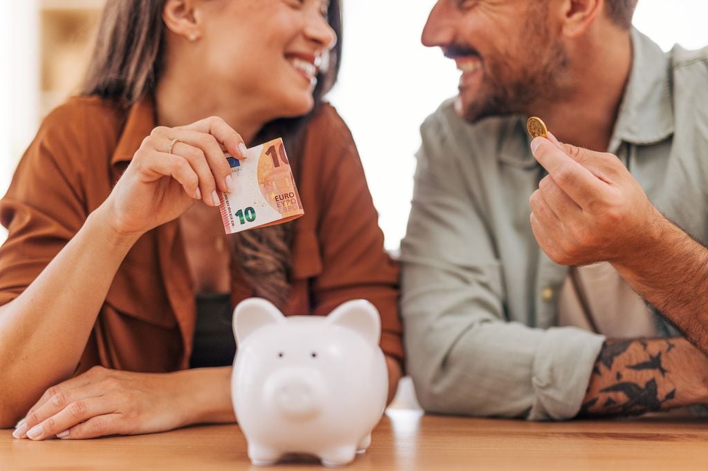 Pareja joven sonriente guardando dinero en una hucha para ahorrar e invertir en su futuro