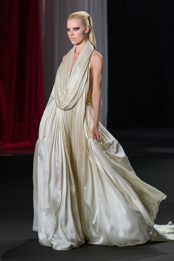Modelo en pasarela con vestido blanco drapeado y maquillaje marcado.