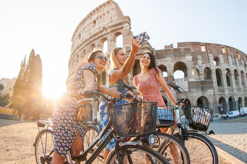 tres chicas sonrientes en bicicleta haciéndose un selfie junto al Coliseo