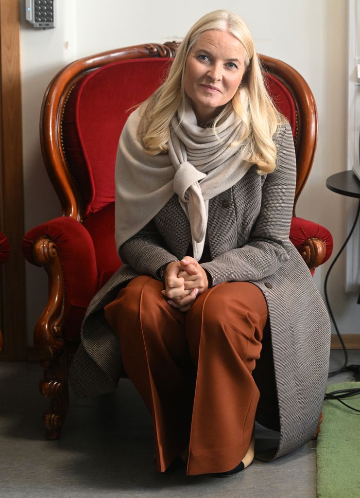 Plano entero de Mette-Marit sentada con abrigo largo marrón y pantalón teja