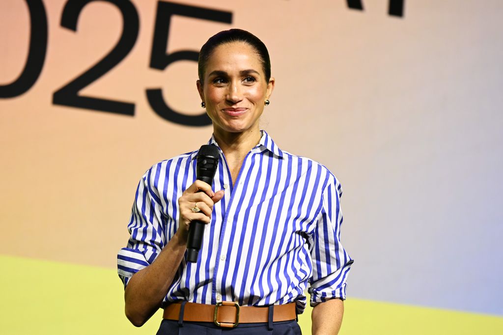 Meghan, Duquesa de Sussex, en camisa azul y blanca a rayas, sostiene un micrófono y sonríe.