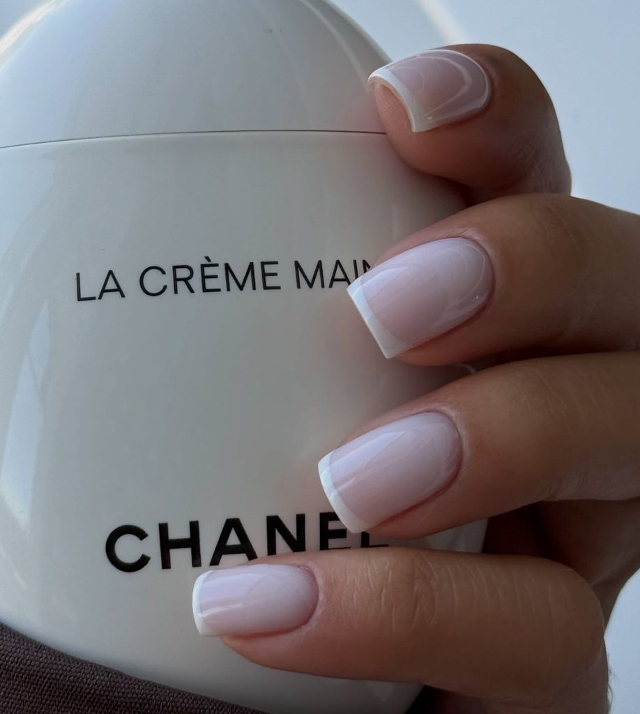 Manos con manicura clara agarrando una crema de manos de Chanel