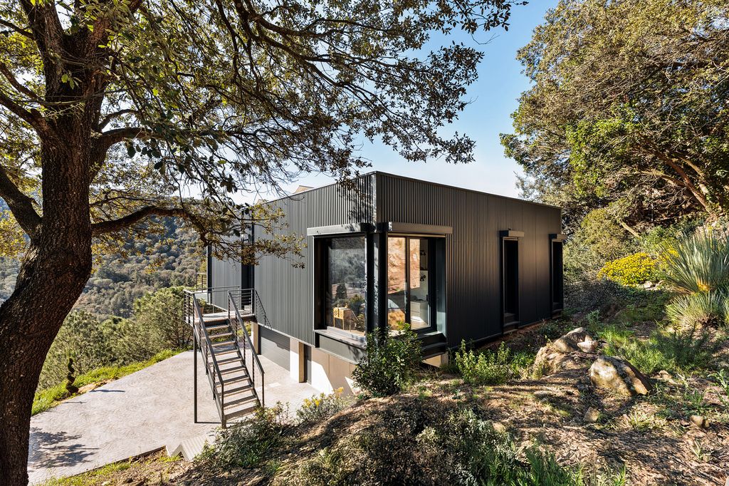 Casa con fachada negra en medio del bosque, ventanal, escalera, árboles