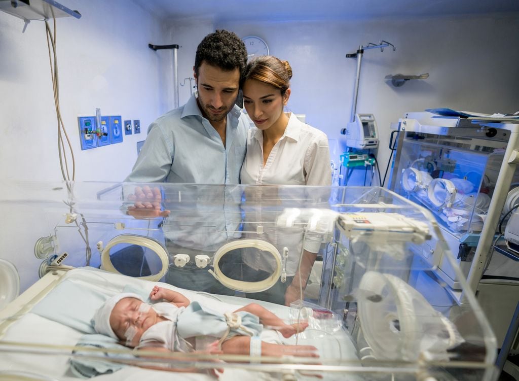 Hombre abrazando a su pareja en la unidad de cuidados intensivos neonatales mientras ambos miran a su hijo recién nacido con amor y preocupación.