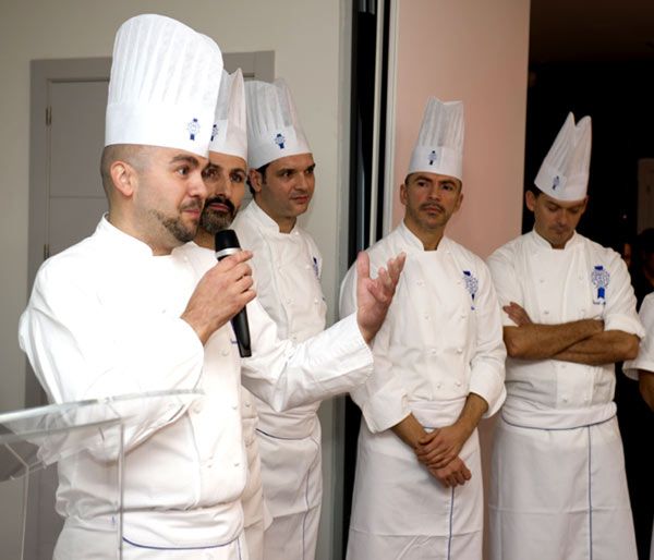 Cinco chefs en uniforme blanco, uno habla en un podio con micrófono.
