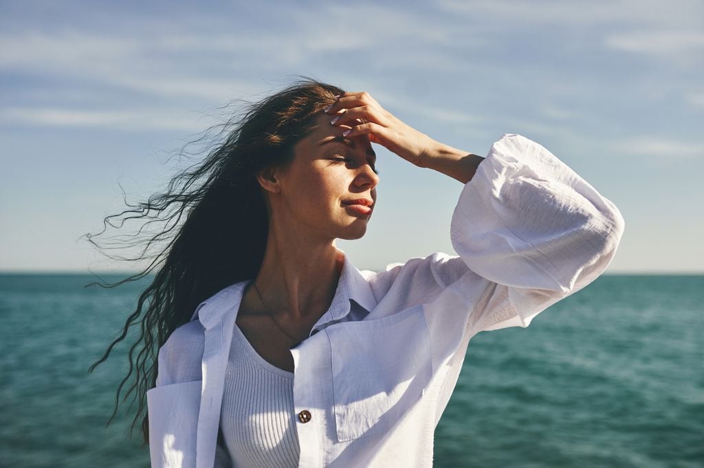 Mujer morena junto al mar con los ojos cerrados, el cabello al viento y la mano en su frente