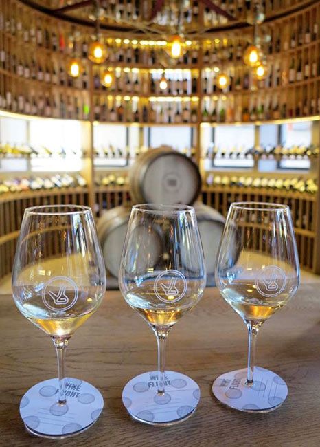 Tres copas de vino en una bodega, con barricas y estantes de vino al fondo.