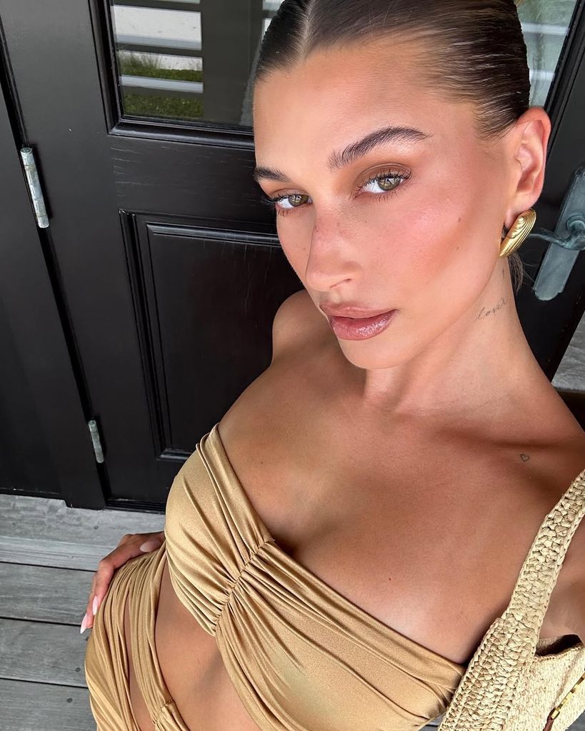 Hailey Bieber con maquillaje efecto glow y piel luminosa, posando con vestido dorado y recogido pulido.
