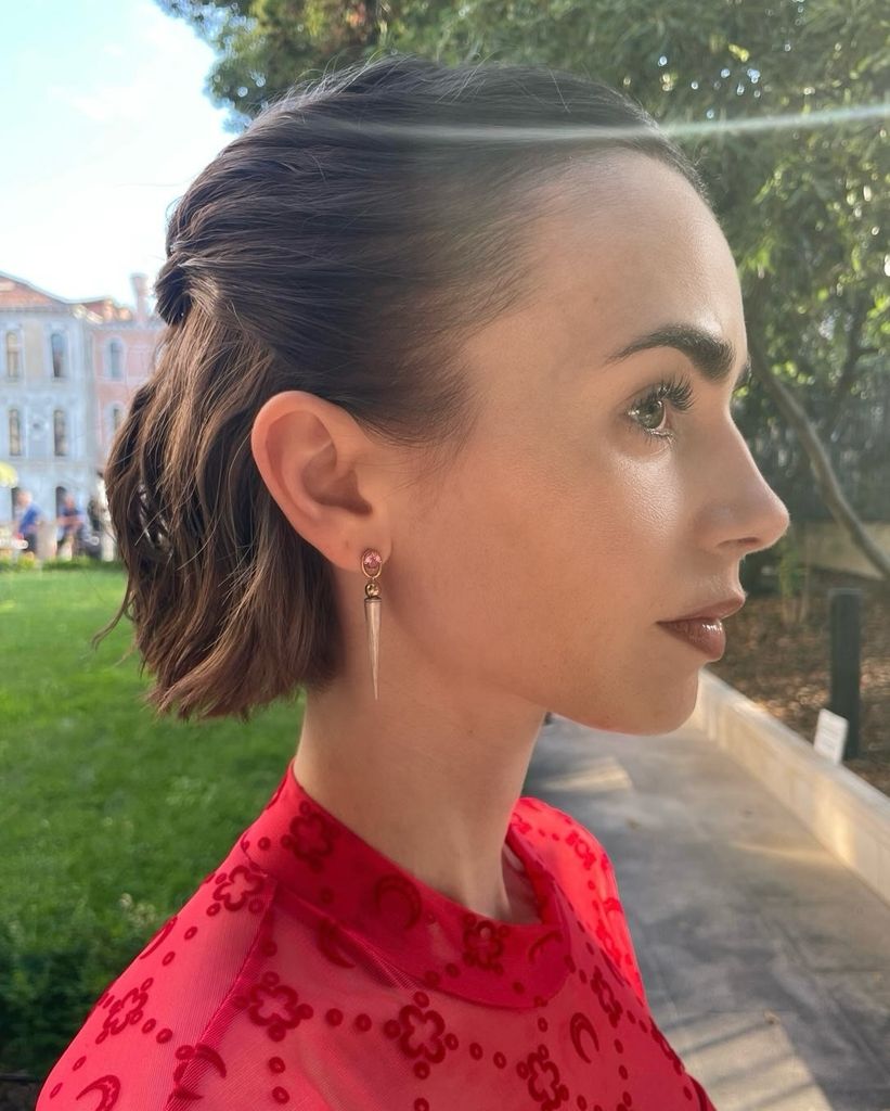 Primer plano lateral del rostro de Lily Collins con peinado recogido hacia atrás, mostrando su pendiente dorado y piel luminosa sin maquillaje.