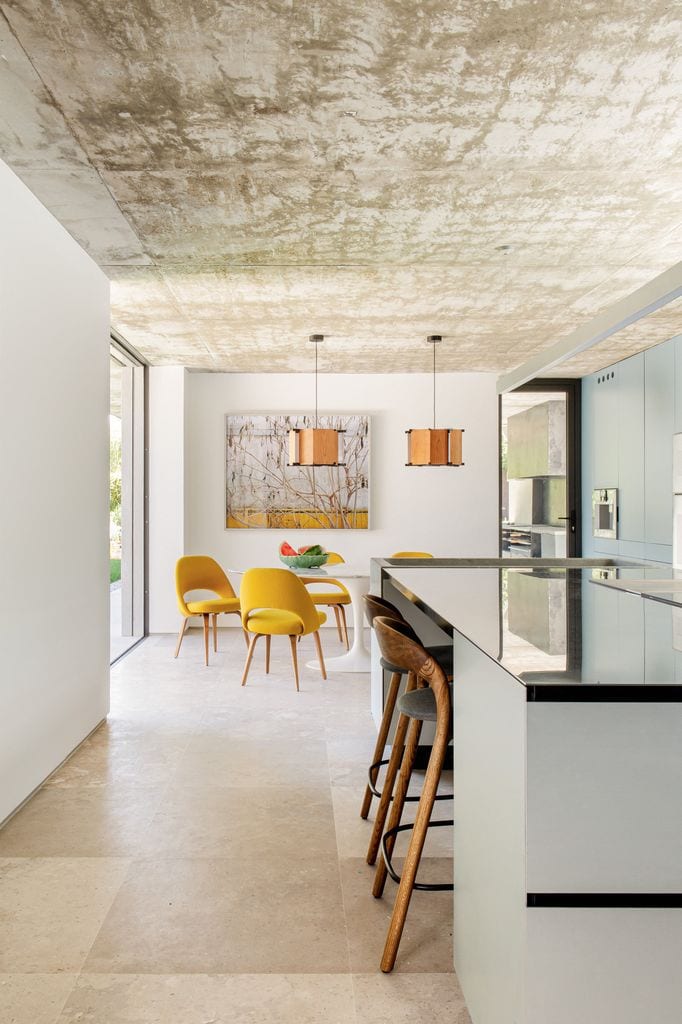 Cocina diseñada por el estudio ÁBATON. En primer plano barra de desayunos alta con taburetes. Al fondo, mesa de comedor con sillas tapizadas en amarillo.