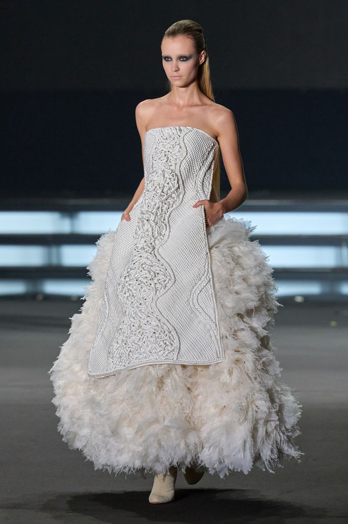 Modelo en pasarela con vestido blanco de plumas y diseño tejido.
