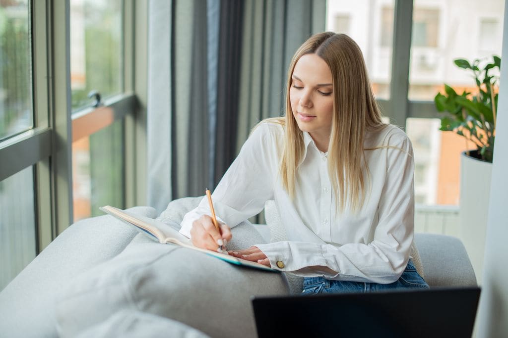 mujer escribiendo en un cuaderno en su casa, junto a una ventana