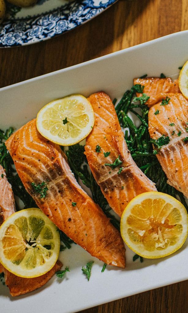 Top down image of four salmon darns on a white long oval serving ceramic plate on a wooden table. It rests on a bed of samphire. Lemon slices garnish the fish.