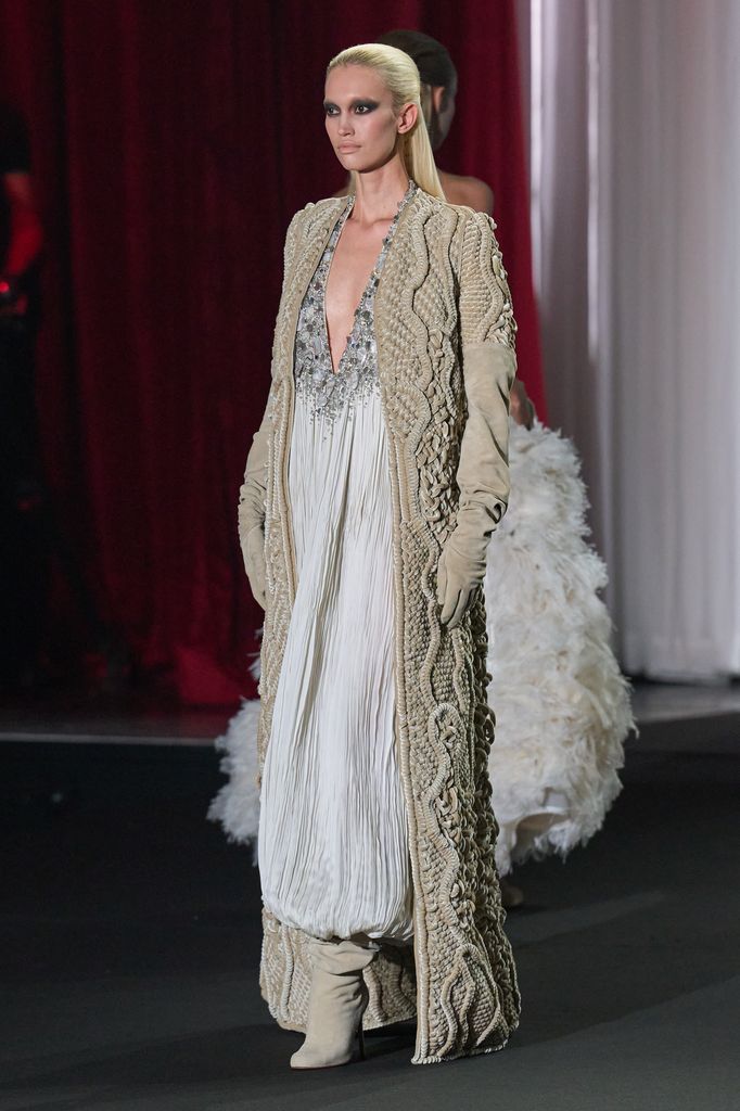 Modelo rubia en pasarela con vestido blanco y capa beige, siguiendo a otra modelo.