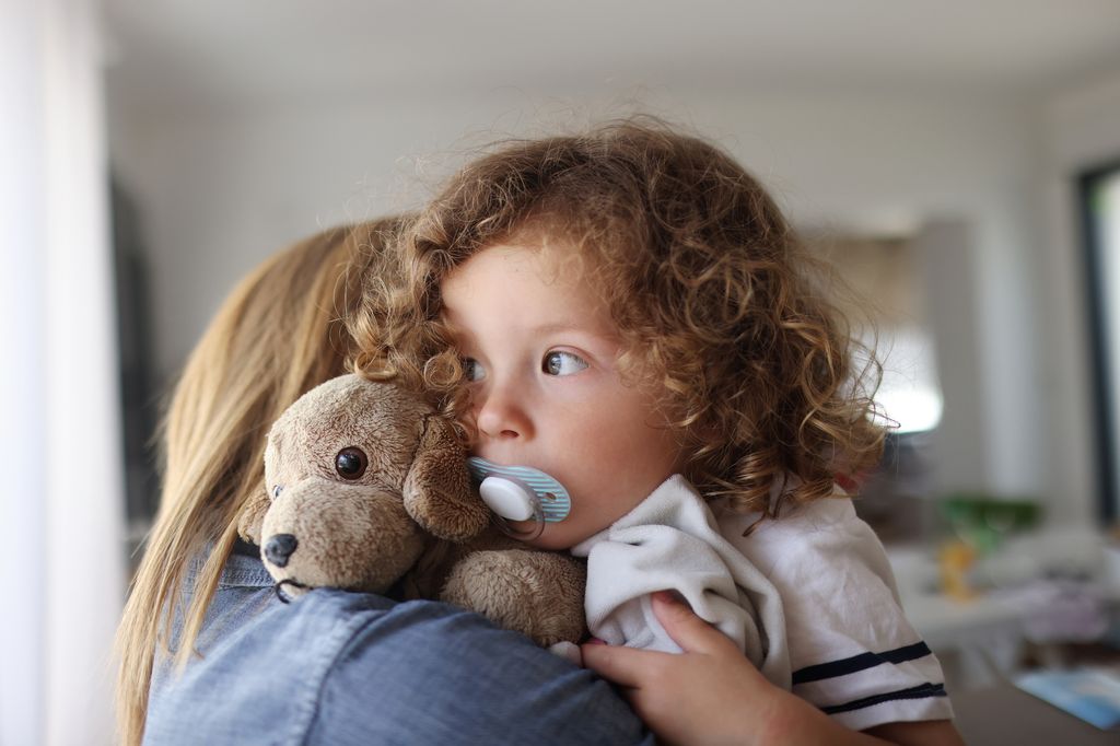 Niña de dos años con chupete y oso de peluche en brazos de su madre
