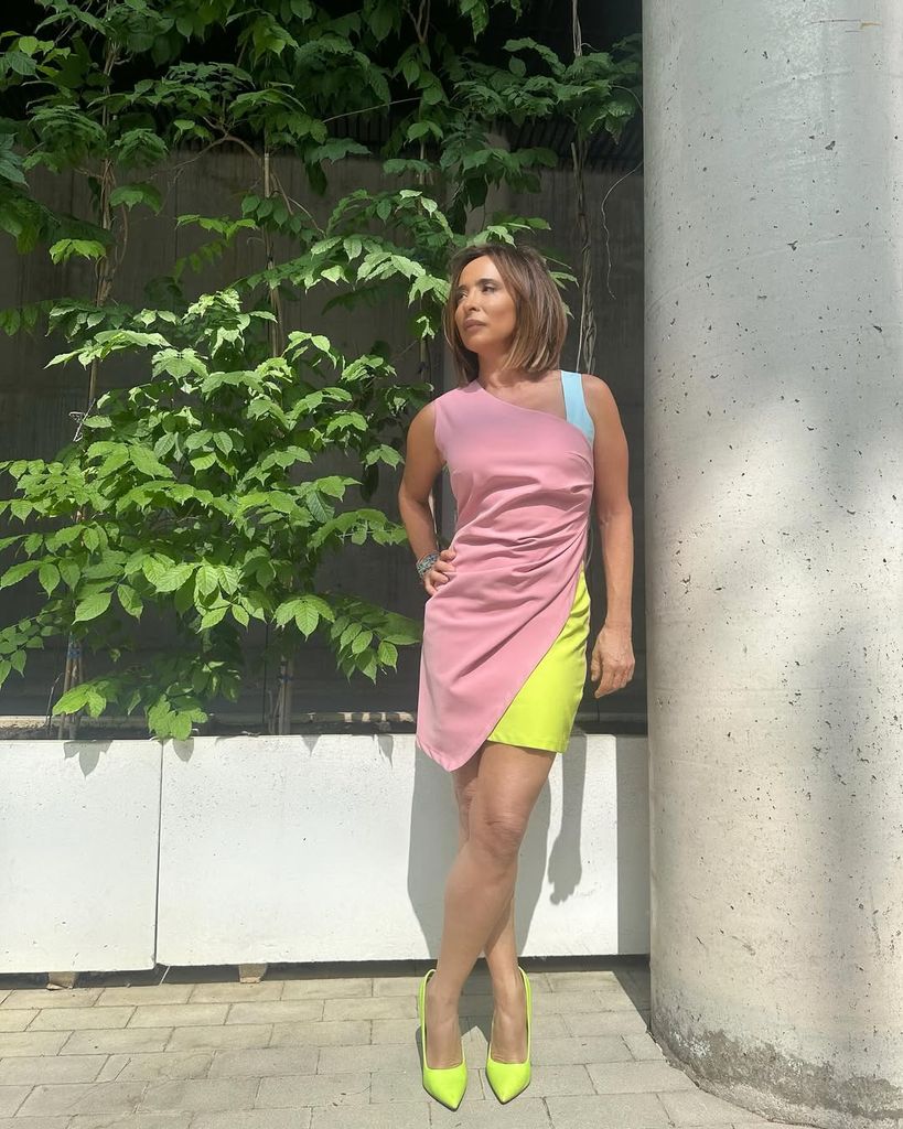 Mujer con cabello corto, en vestido rosa y amarillo, tacones verdes, junto a columna y plantas.