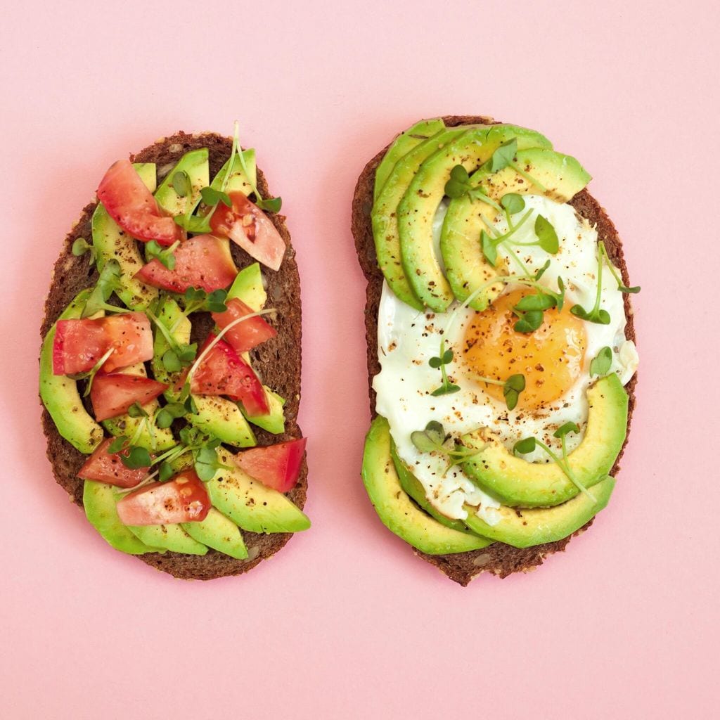 Toasts of dark bread with avocado slices, red tomatoes, fried egg and microgreen. Top view with pink background.
