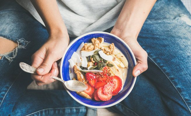 Persona con jeans y camiseta gris sosteniendo un tazón de comida con cuchara.