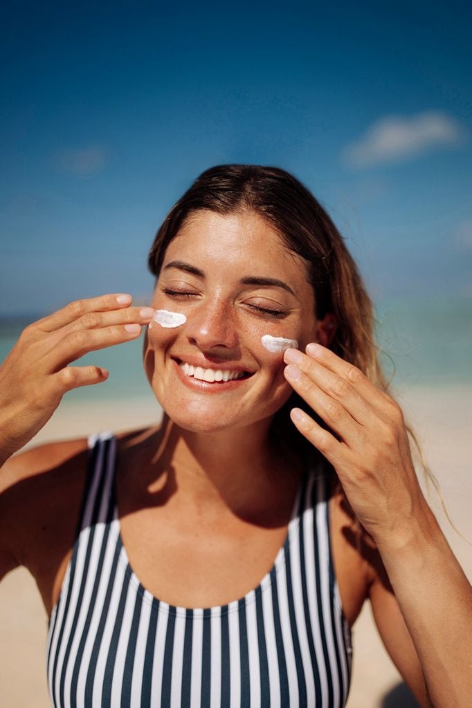 Mujer sonriente en la playa aplicando protector solar en la cara con bañador de rayas
