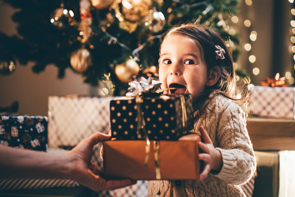 Niña pequeña recibiendo paquetes de Navidad al lado del árbol con cara de sorpresa.
