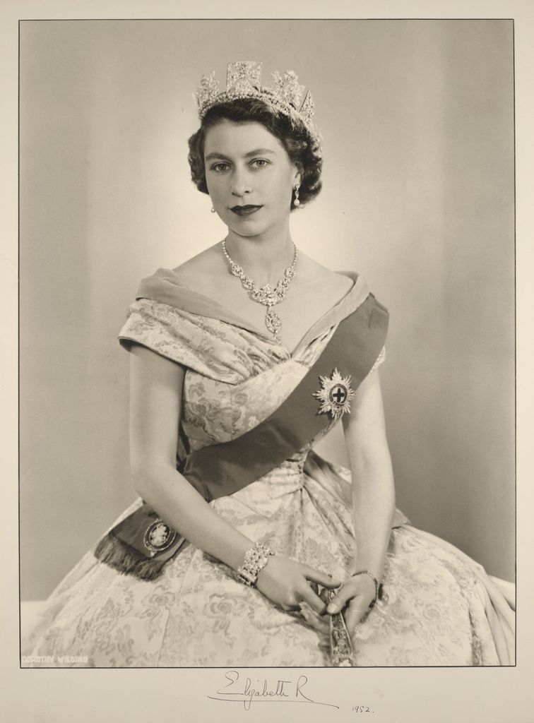 Retrato fotográfico de Isabel II sentada de frente y sosteniendo un abanico cerrado en su regazo. Viste un vestido de noche de satén de Norman Hartnell y la estrella y el emblema de la Orden de la Jarretera en una banda, así como la Diadema de Diamantes de Estado o de Jorge IV, un collar de diamantes que le obsequió el Nizam de Hyderabad y una pulsera de diamantes que fue un regalo de bodas de Su Alteza Real el Duque de Edimburgo. Firmado por la Reina en el soporte y fechado en 1952. Firma del estudio de Dorothy Wilding en blanco en la esquina inferior izquierda.