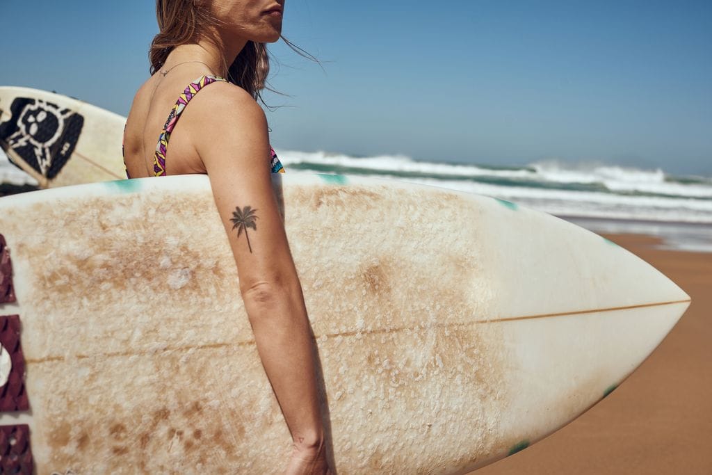 mujer con una tabla de surf y un tatuaje en el brazo