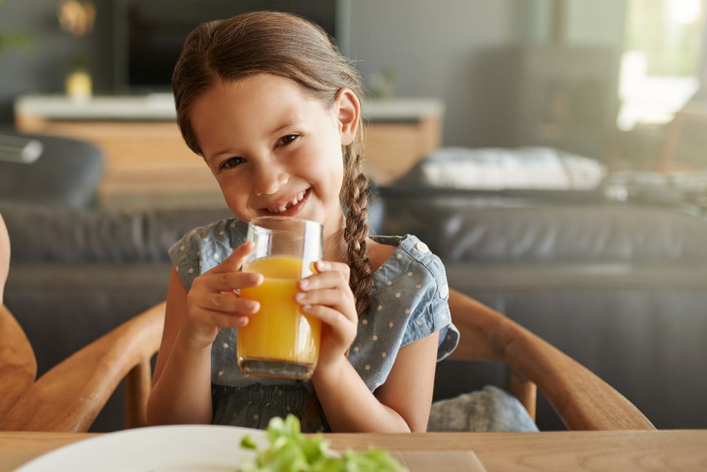Niña feliz con trenzas, bebiendo jugo naranja en casa.