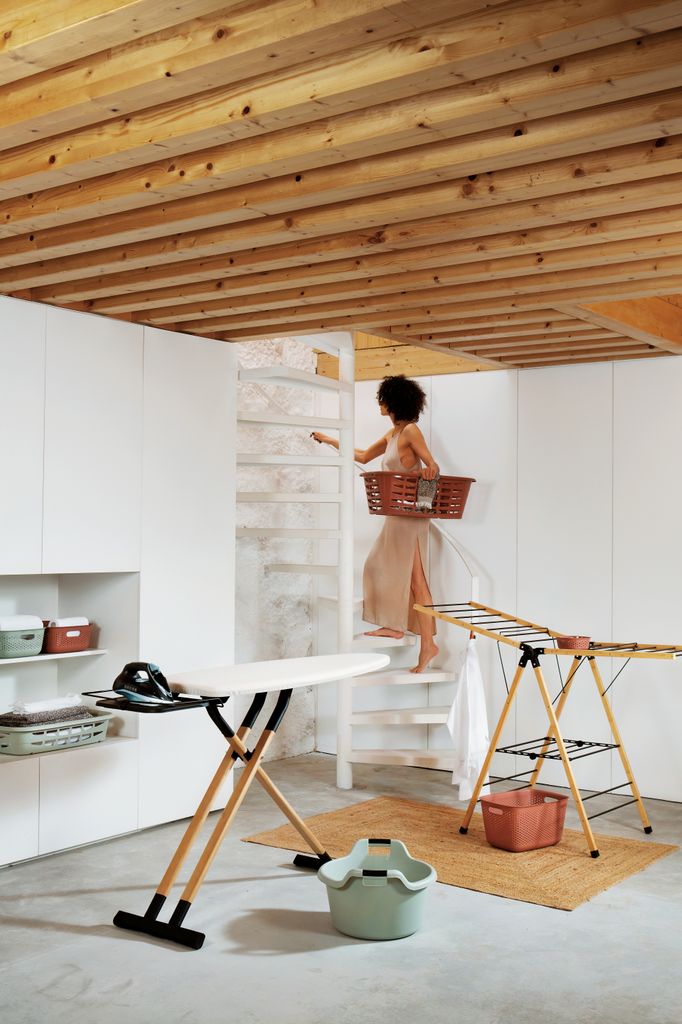 Mujer en sótano con cesta de ropa, tabla de planchar, tendedero portátil, cestos y armario con baldas 