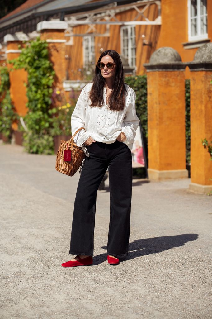 Mujer con cabello largo, camisa blanca, jeans negros y zapatos rojos, posando en una calle.