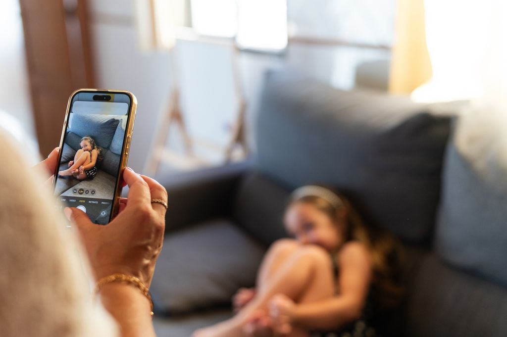 Madre grabando a su hija pequeña sentada en el sofá