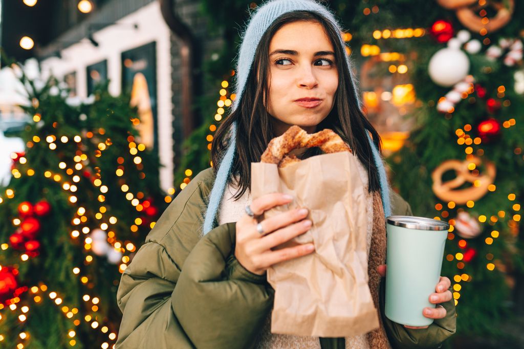 Mujer comiendo un pretzel con queso y un café en termo en un mercadillo navideño.