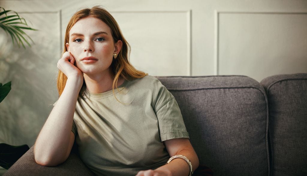Mujer con cabello rubio, camiseta verde y pulsera plateada sentada en sofá gris.