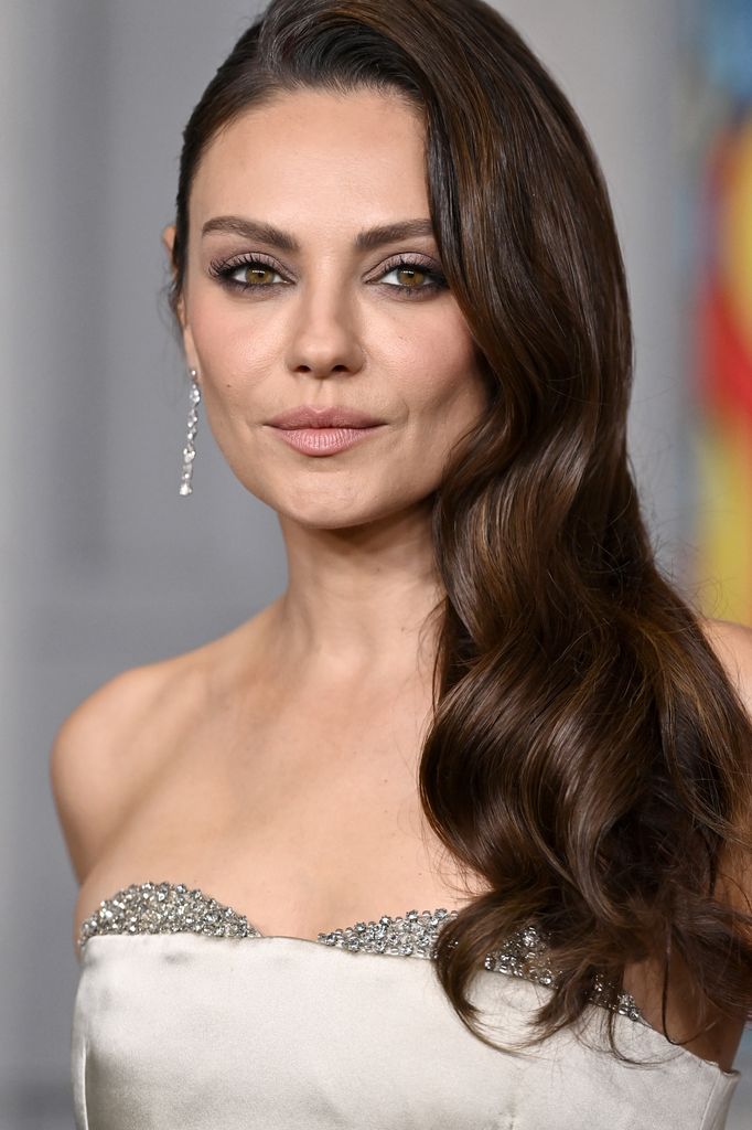 Mila Kunis en la premiére de Wake Up Dead Man: A Knives Out Mystery con el pelo suelto ondulado y vestido blanco palabra de honor