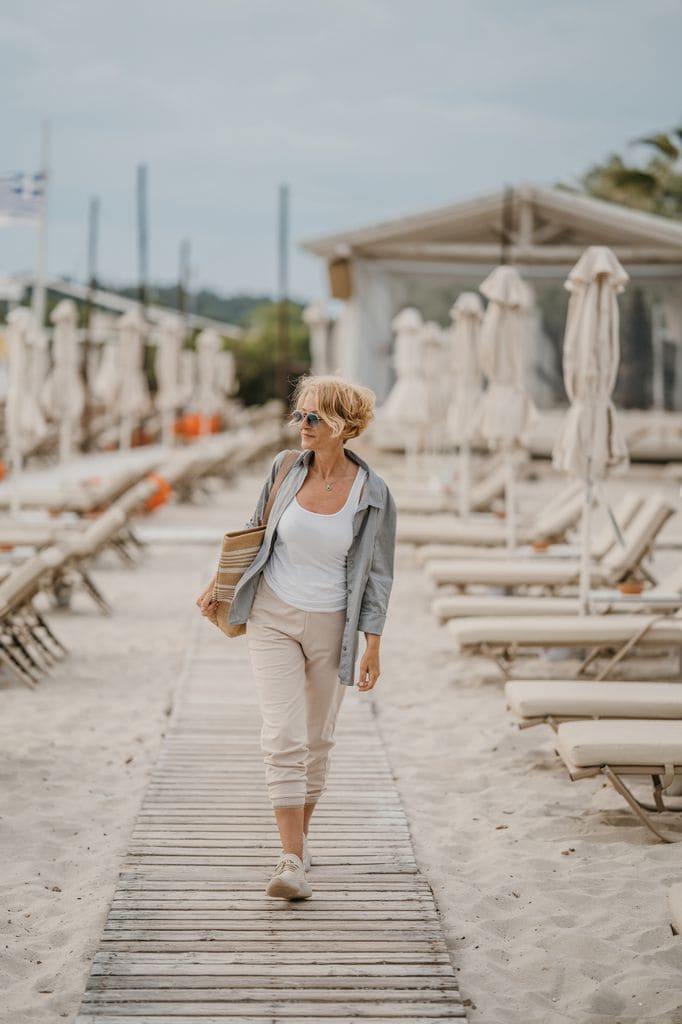 Mujer madura caminando por la playa con zapatillas, pantalones beis, camiseta, camisa y bolso grande