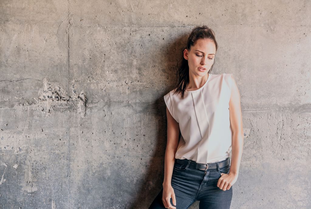 Mujer con cabello oscuro, camisa blanca y jeans, posando contra pared de concreto.