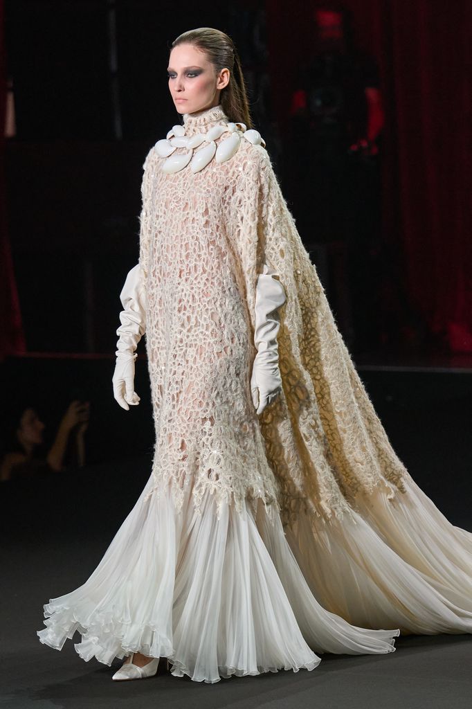 Modelo caminando en pasarela con vestido blanco de encaje y guantes blancos.