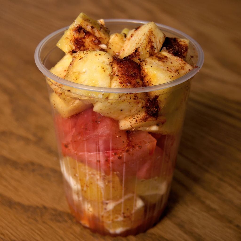 Plastic cup of fruta picada [chopped pineapple, watermelon and orange], seasoned with lime juice and chilli flakes, on a wooden table