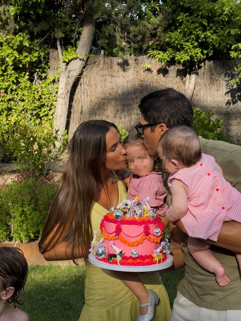 Un hombre y una mujer besan a un bebé en un jardín con pastel.